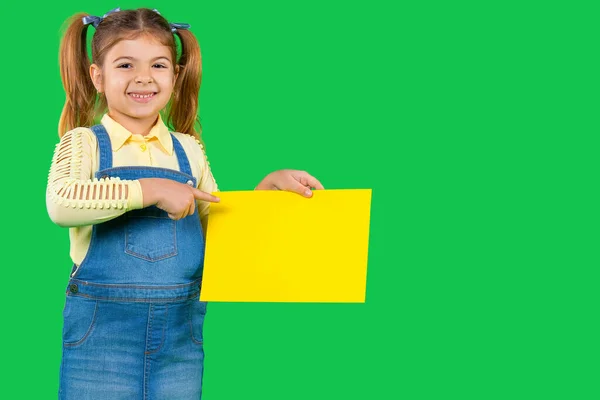 Sorrindo menina pré-escolar segurando um banner amarelo para o seu anúncio em um fundo verde com espaço lateral em branco. — Fotografia de Stock