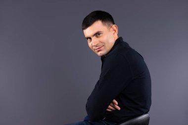 Gri arka planda deri koltuklu siyah gömlekli şık bir adam kameraya bakar. Profil fotoğrafı.