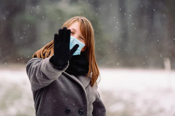 Menina caucasiana em uma máscara azul descartável médica mostra parada na câmera no fundo nevado floresta de inverno. — Fotografia de Stock