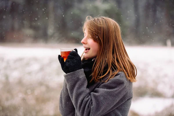 Congelado, mas satisfeito bonito menina loira com uma xícara de chá quente no fundo de uma paisagem de floresta de inverno gelada — Fotografia de Stock