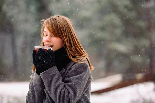 Uma menina satisfeita com os olhos fechados goza do aroma de chá quente no fundo de uma floresta e pequenos flocos de neve caindo. — Fotografia de Stock