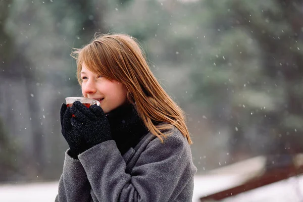 From the cold, a frozen girl holds a cup of hot tea, enjoys the aroma and looks to the side against the background of a winter forest.