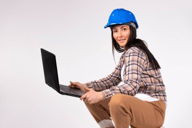 Mavi kasklı bir kadın beyaz arka planda dizüstü bilgisayarla poz veriyor ve kameraya gülümsüyor..