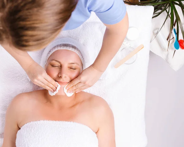L'estetista pulisce il viso di una donna avvolta in un asciugamano bianco con batuffoli di cotone in un salone di bellezza. Idratante e ringiovanente della pelle. — Foto Stock