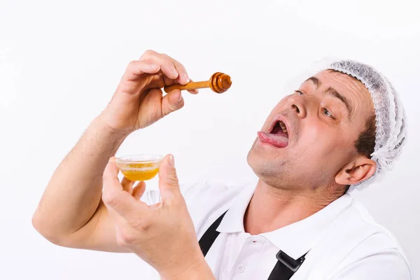 En kock med ett lustigt uttryck försöker honung från en skål på en vit bakgrund. — Stockfoto