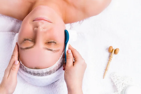 Retrato de uma mulher que está fazendo massagem facial com um massageador cosmético deitado em uma toalha branca. — Fotografia de Stock