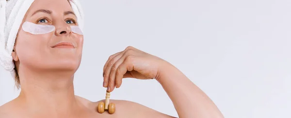 Banner de formato largo. Mujer anciana sonriente con parches limpios de piel y ojos, mirando hacia arriba, sostenga un masajeador de piel de madera de rodillo. Masaje automático sobre fondo blanco y espacio en blanco lateral. —  Fotos de Stock