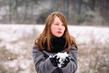 Düşünceli bir kız elinde bir kar küresi tutuyor ve kışın karla kaplı ormanın arka planına bakıyor..