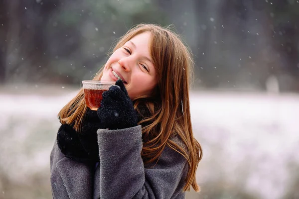 Портрет девушки с чашкой горячего чая и счастливой улыбкой, смотрящей в камеру на фоне леса. — стоковое фото