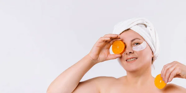 Bannière... format long. Belle femme âgée souriante avec des patchs pour les yeux, regardant vers la caméra, tenant une tranche d'orange fraîche. Skinimalism soins, spa, beauté naturelle et concept de cosmétologie. — Photo