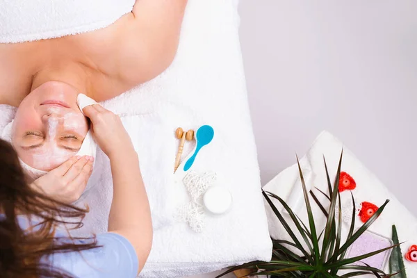 A beautician wipes a mask off a adult womans face, in a salon with green flowers and scented candles in background. Cosmetologist makes a facial skin care procedure. — 图库照片