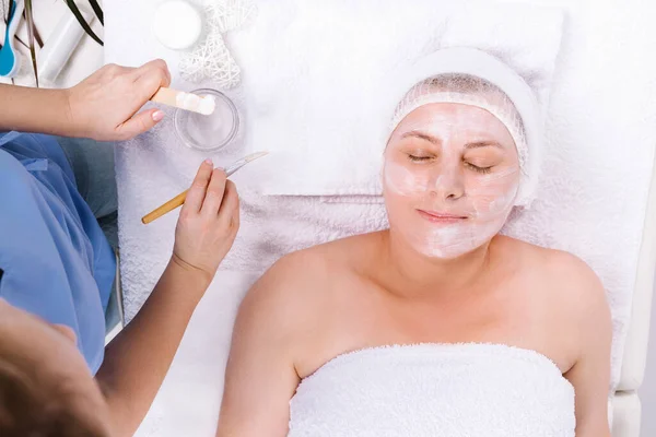 The female beautician uses a brush and a spatula to apply a cream mask to the senior clients face, which lies on a white towel. Senior spa and beauty — Stock fotografie
