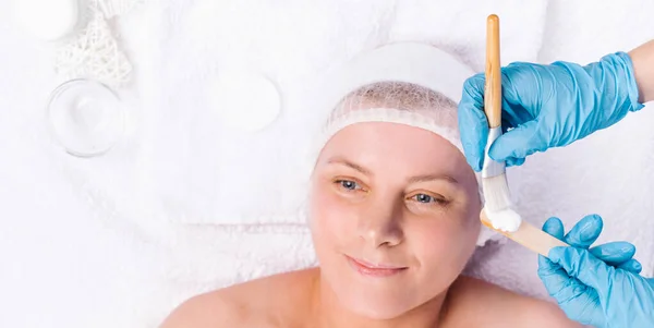 Banner. Eine Frau mit einem kosmetischen Kopfverband liegt in einem Salon auf einem weißen Handtuch. Die Hände der Kosmetikerin bereiten die Gesichtsmaske mit Kosmetikpinsel und Holzspachtel vor. — Stockfoto