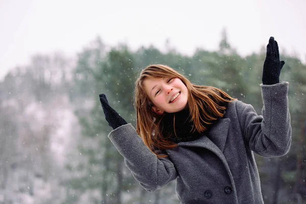 Menina feliz em um casaco cinza e um lenço quente posando com os braços estendidos para os lados no fundo de uma floresta nevada. — Fotografia de Stock