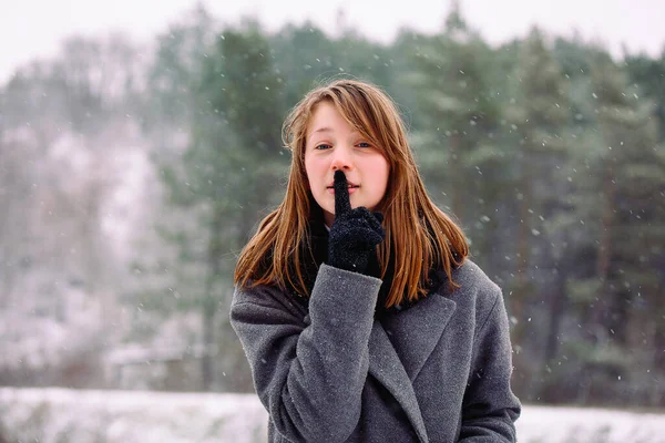 Vit flicka i grå rock och i svarta handskar visar sitt pekfinger tyst vid kameran mot bakgrund av en vinterskog. — Stockfoto