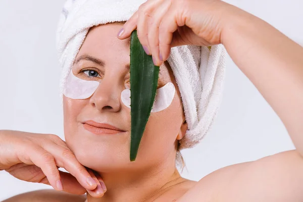 Retrato de anciana caucásica con piel clara, parches cosméticos para los ojos y hoja de aloe vera frente a la cara, cerrando un ojo. Fondo blanco. —  Fotos de Stock