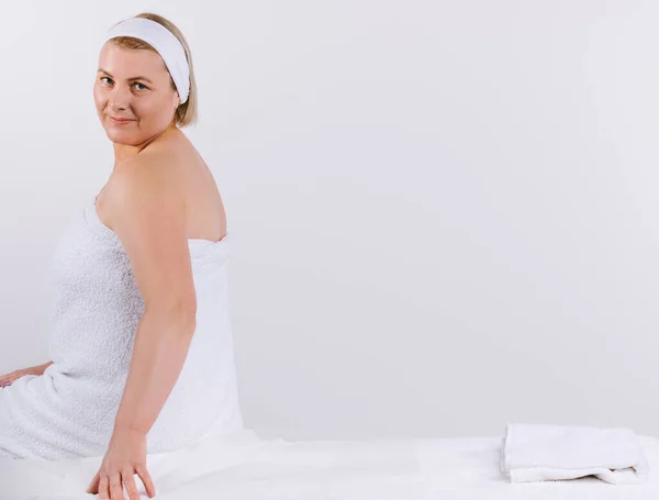 Senior woman, wrapped in a white towel, sits on a massage table before the procedure and looks cute at the camera. White background with empty space for advertising. — Stock Photo, Image