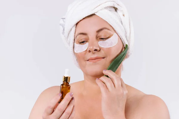 Seniorin mit Einweg-Augenklappen, Lifting-Serum und Aloe-Vera-Blatt schaut weg. Weißer Hintergrund. — Stockfoto