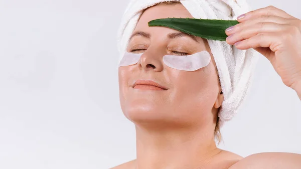 Ein vergrößertes Foto einer älteren Frau mit heller Haut, kosmetischen Augenklappen und Aloe-Vera-Blatt. Weißer Hintergrund. — Stockfoto