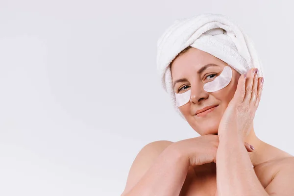 Zadowolona starsza kobieta pozująca na białym tle z ręcznikiem na głowie i ochraniaczami pod oczami. Boczna przestrzeń reklamowa. — Zdjęcie stockowe