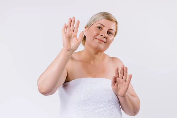 Une femme âgée enveloppée dans une serviette blanche avec un sourire pose et montre les mains douces saines et hydratées sur un fond blanc. — Photo