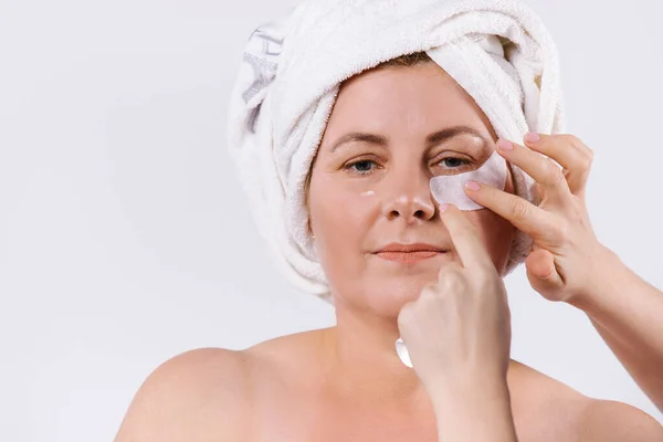 Elderly woman with white towel on her face and hands of beautician applying bandages under her eyes. White background. — 스톡 사진