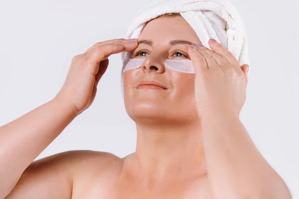 Powiększenie zdjęcia starszej kobiety o jasnej skórze z białym ręcznikiem na głowie i plamami pod oczami patrzącymi na białe tło. Chuda i promienna skóra. — Zdjęcie stockowe