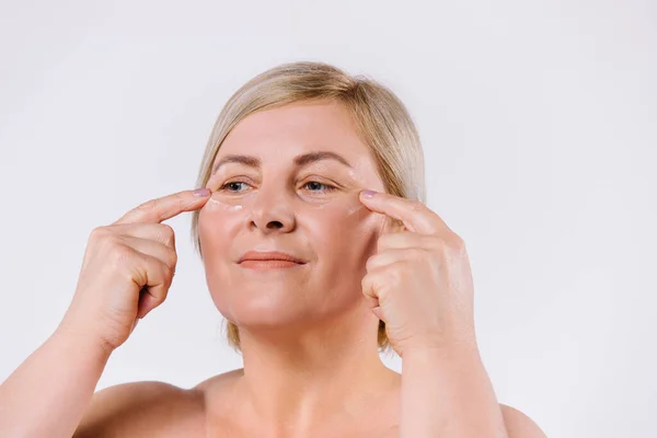 Uma mulher mais velha aplica um creme revitalizante sob os olhos com os dedos indicadores. Foto ampliada. Fundo branco. — Fotografia de Stock