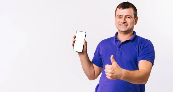 Banner. Deaf senior man shows thumb up while holding a mobile phone, white screen to the camera. White background and blank advertising space. — Stock Photo, Image