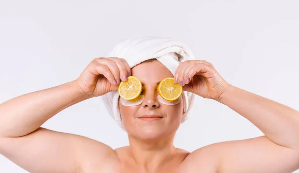 Starší žena s bílým ručníkem na hlavě drží citrónové klíny nad očima. Bílé pozadí. — Stock fotografie