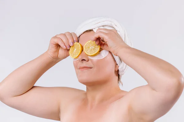 Portrait of a senior woman with a towel on her head, looks to the side and holds lemon slices in front of her eyes. White background. — Stock fotografie