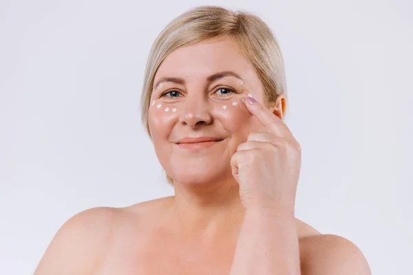 Gözlerinin altında krem maskeli yaşlı sarışın kadın. Mükemmel cilt ve kremi olan, yaşlanmayı önleyen ve kırışık gözlü güzel bir kadın. Güzellik ve mükemmellik kavramı
