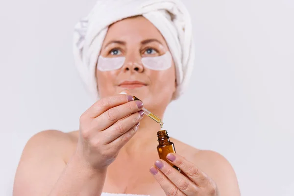 An older woman, looking up, with disposable eye patches and a white towel on her head holds a anti-age serum in front of her. White background. — Stockfoto