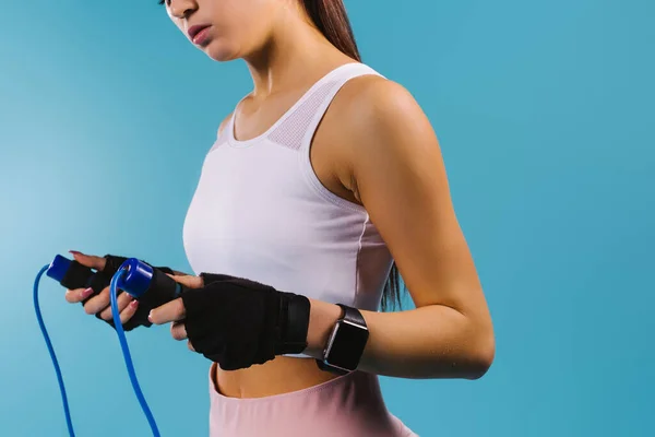 Foto recortada de uma menina esportiva em um top branco, com uma corda e um relógio fitness em um fundo azul. — Fotografia de Stock