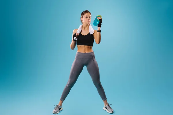 Mladá dívka s atletickou postavou pije proteinový koktejl, zatímco odpočívá mezi cvičeními. Modré pozadí a spousta volného místa pro reklamu. — Stock fotografie