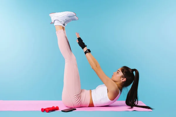 A menina esportiva em uma esteira de fitness faz exercícios da prensa. Fundo azul com espaço vazio. — Fotografia de Stock