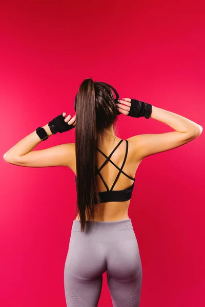 Photo from behind. A brunette with pumped up buttocks listens to music in large headphones on a red background. Sports concept and beautiful figures. — Stock Photo, Image
