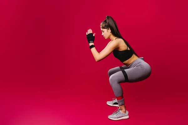 Esmer, spor elbiseli, elastik bandajlı bir kız. Kırmızı arka planda egzersiz yapıyor..