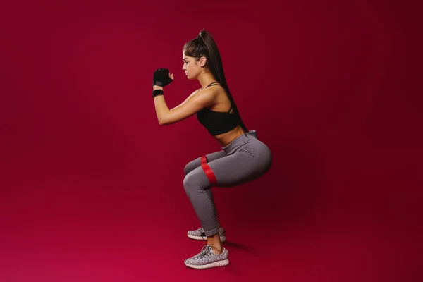Λεπτό κορίτσι σε αθλητικά ρούχα με ελαστικό επίδεσμο κάνει ασκήσεις σε κόκκινο φόντο με κενό χώρο στο πλάι. Η έννοια ενός υγιεινού τρόπου ζωής και μια όμορφη φιγούρα. — Φωτογραφία Αρχείου