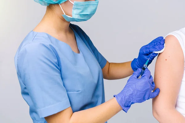 Beskär foto av en läkare i medicinsk uniform och engångsmask sätta covid-19 vaccin på en patient underarm mot en grå bakgrund. — Stockfoto