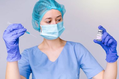 Tıbbi maskeli ve steril eldivenli genç bir kadın doktor ellerinde bir şırınga tutuyor ve gri arka planda 19 aşılı bir ampule bakıyor..