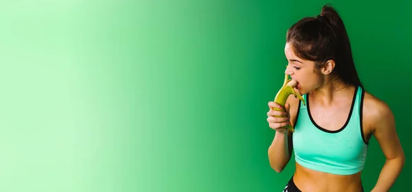 Банер, довгий формат. Концепція здорової їжі. Фітнес дівчина в спортивному одязі на зеленому тлі їсть банан за міцність та енергію. Концепція здорової їжі . — стокове фото