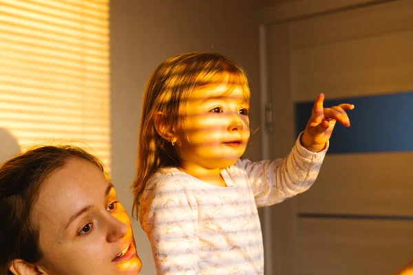 Foto recortada de mamá y niña sentada en casa durante una pandemia. La niña señala con el dedo al sol que brilla fuera de la ventana. — Foto de Stock