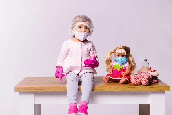 Tıbbi maskeli küçük bir kız ve beyaz arka planda oyuncakları olan pembe lateks eldivenler. Pediatri kliniği ve genç hastalara yaklaşım.