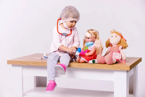 Una niña pequeña, vestida con una bata médica blanca y una máscara médica, interpreta a un médico y trata sus juguetes para varias enfermedades sobre un fondo blanco con espacio lateral. — Foto de Stock