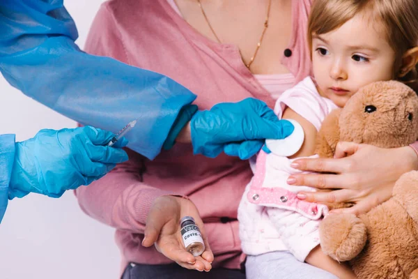 Fotografía recortada. Mamá tiene una hija pequeña con juguete durante la vacunación covid 19. Niña caucásica niño mirada calma en la jeringa — Foto de Stock