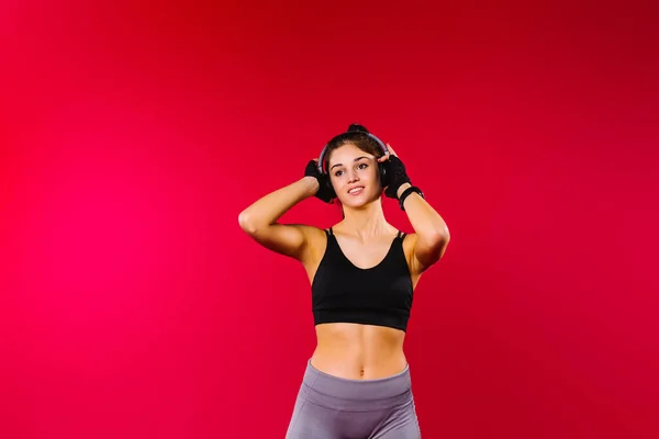 Caucásico chica deportiva en un top deportivo negro y grandes auriculares se ve a un lado en un fondo rojo con espacio lateral vacío. —  Fotos de Stock