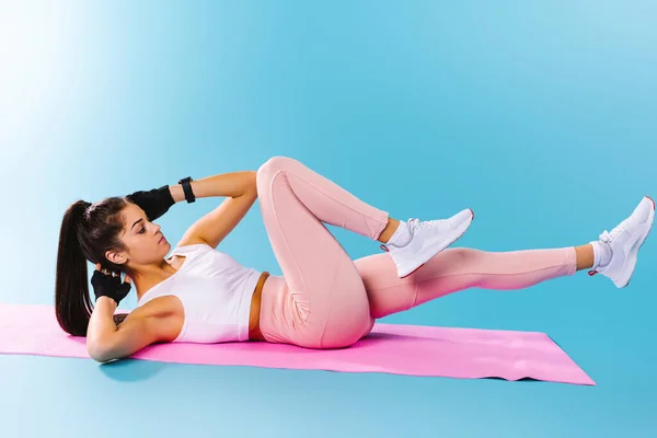 Beyaz spor bluzlu beyaz esmer kız ve yoga minderinin üstünde abdominal egzersizler yapan tayt. Mavi arkaplan.