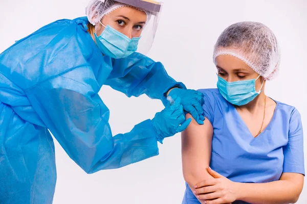 Koruyucu ekipman giyen, tıbbi maske takan bir hemşire, genç bir kadını covid 19 aşısı yapar ve kameraya bakar. Beyaz arkaplan.