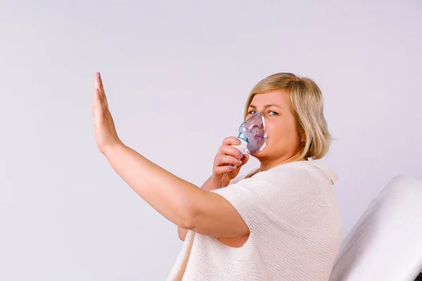 Старша біла жінка вдихає маскою туманності в домашніх умовах при коронавірусному захворюванні, показуючи знак стоп на сірому фоні . — стокове фото